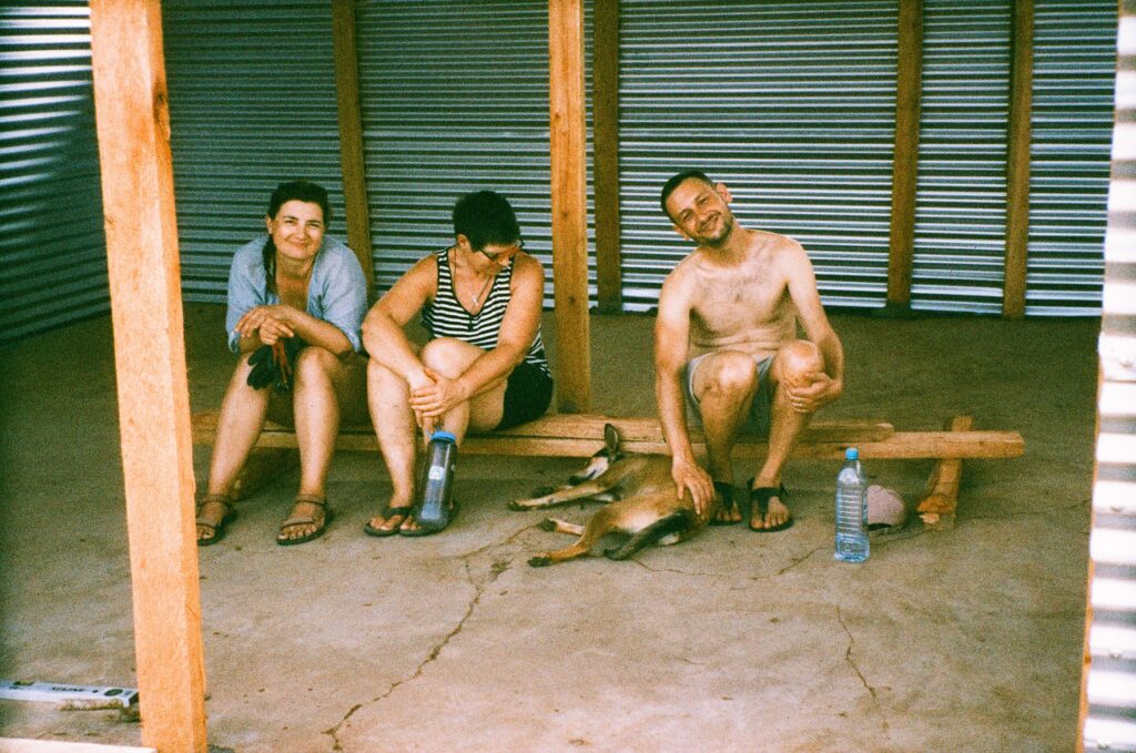 Tři lidé sedí v plechové boudě, dvě ženy a muž bez trička, který hladí psa.